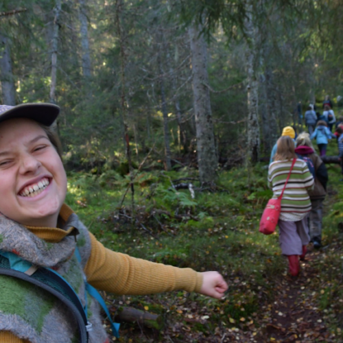 Skog. Jente med caps, turklær og ryggsekk snur seg glisende mot kamera, ungdommer går på stien foran.